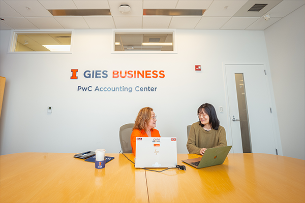 Staff and Student Talking at Table in front of PwC Accounting Center and Gies Business Sign